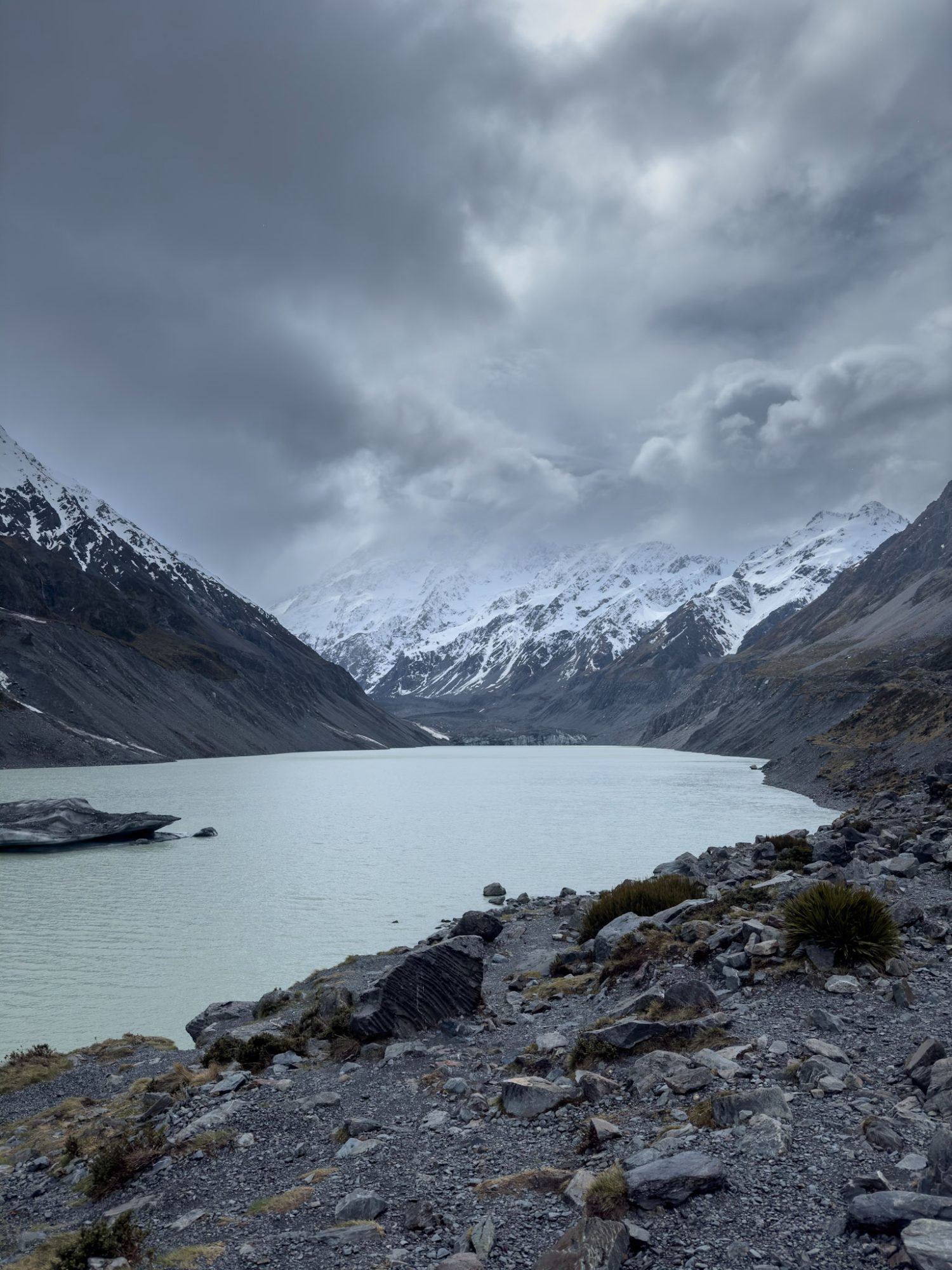 A pale blue lake beneath snow-covered mountains and a rocky shore