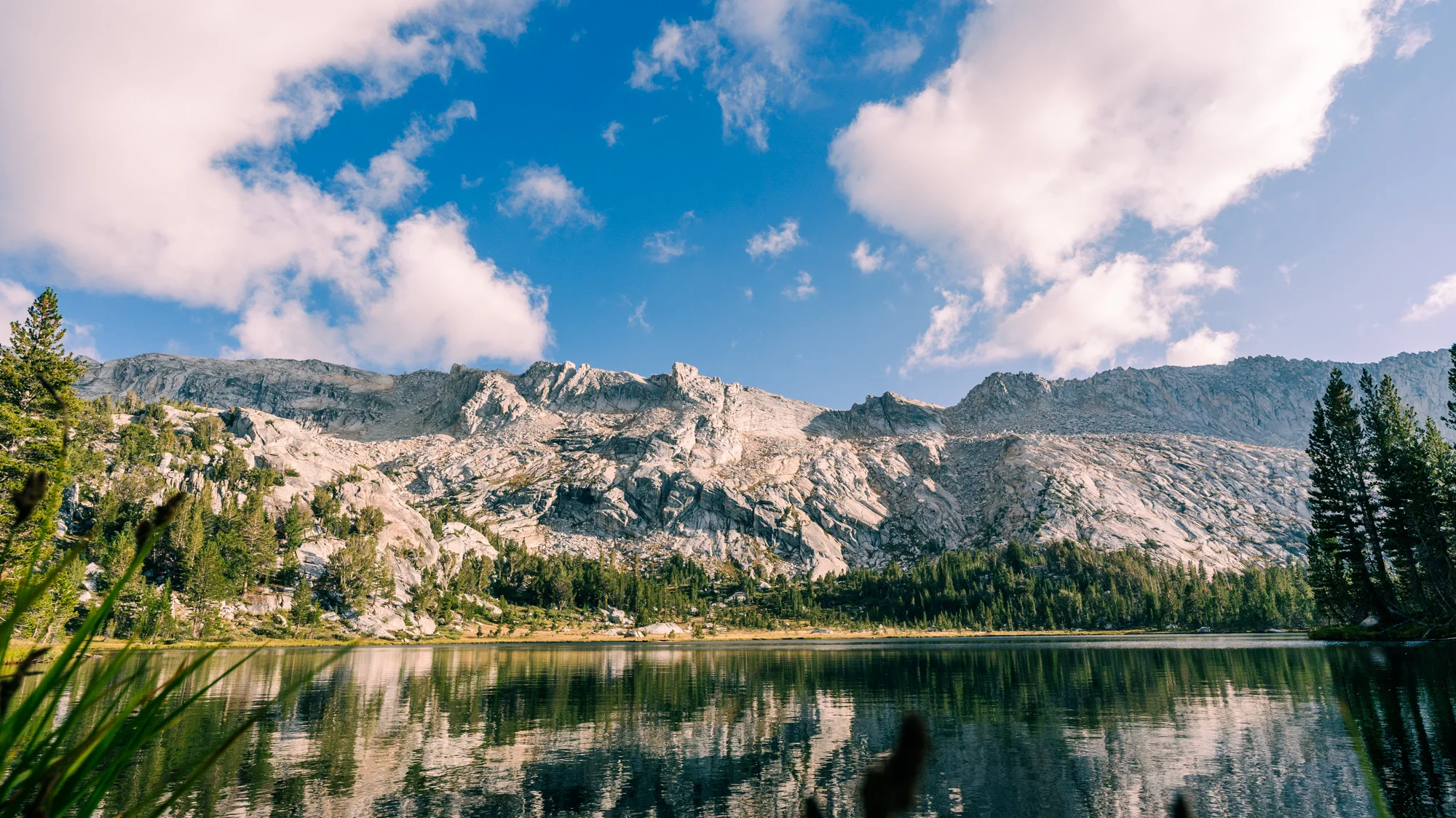 Granite mountains and evergreen trees reflected in a blue lake beneath pink-tinged clouds.
