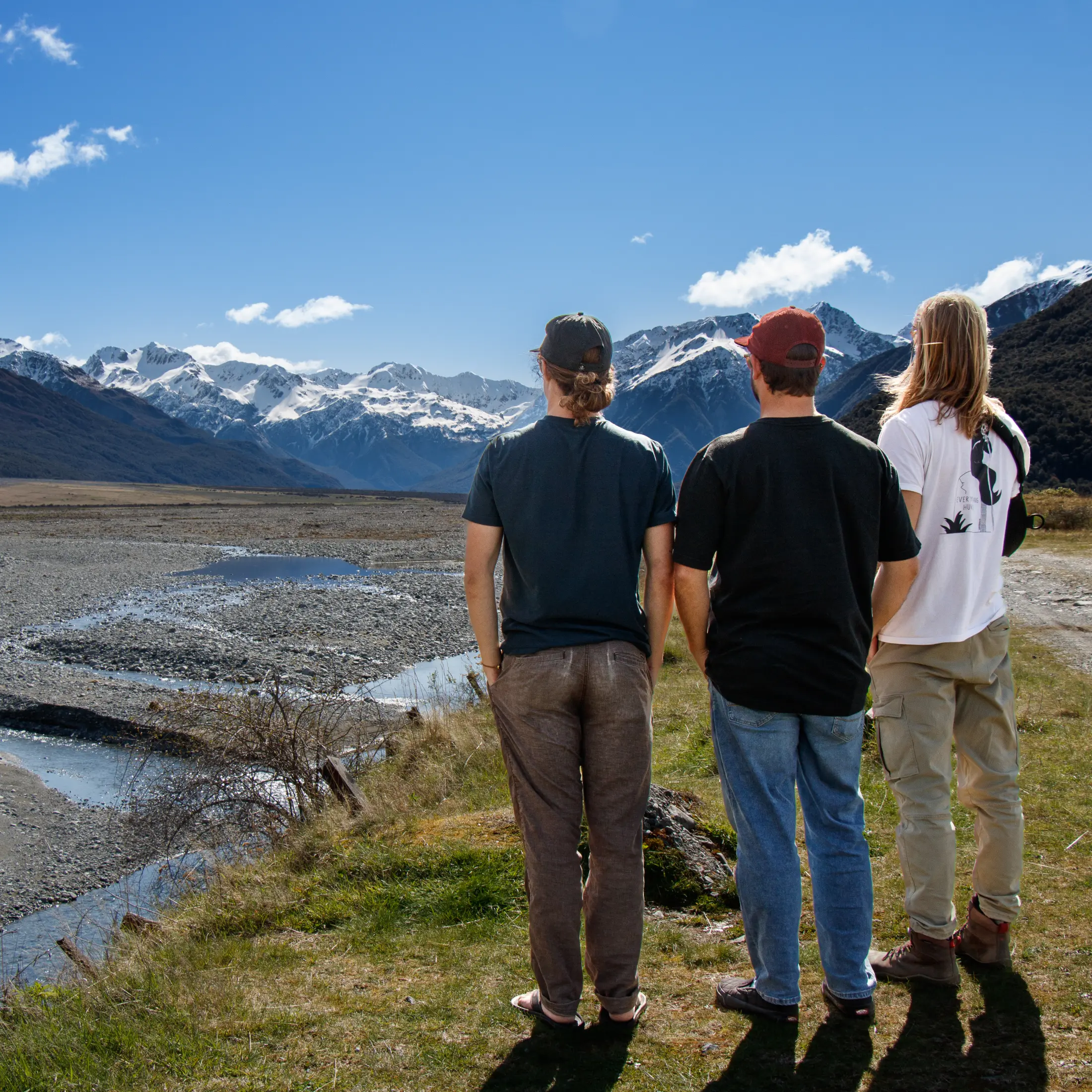 Three friends with their backs to the camera, looking across a braided river toward snow-capped mountains.