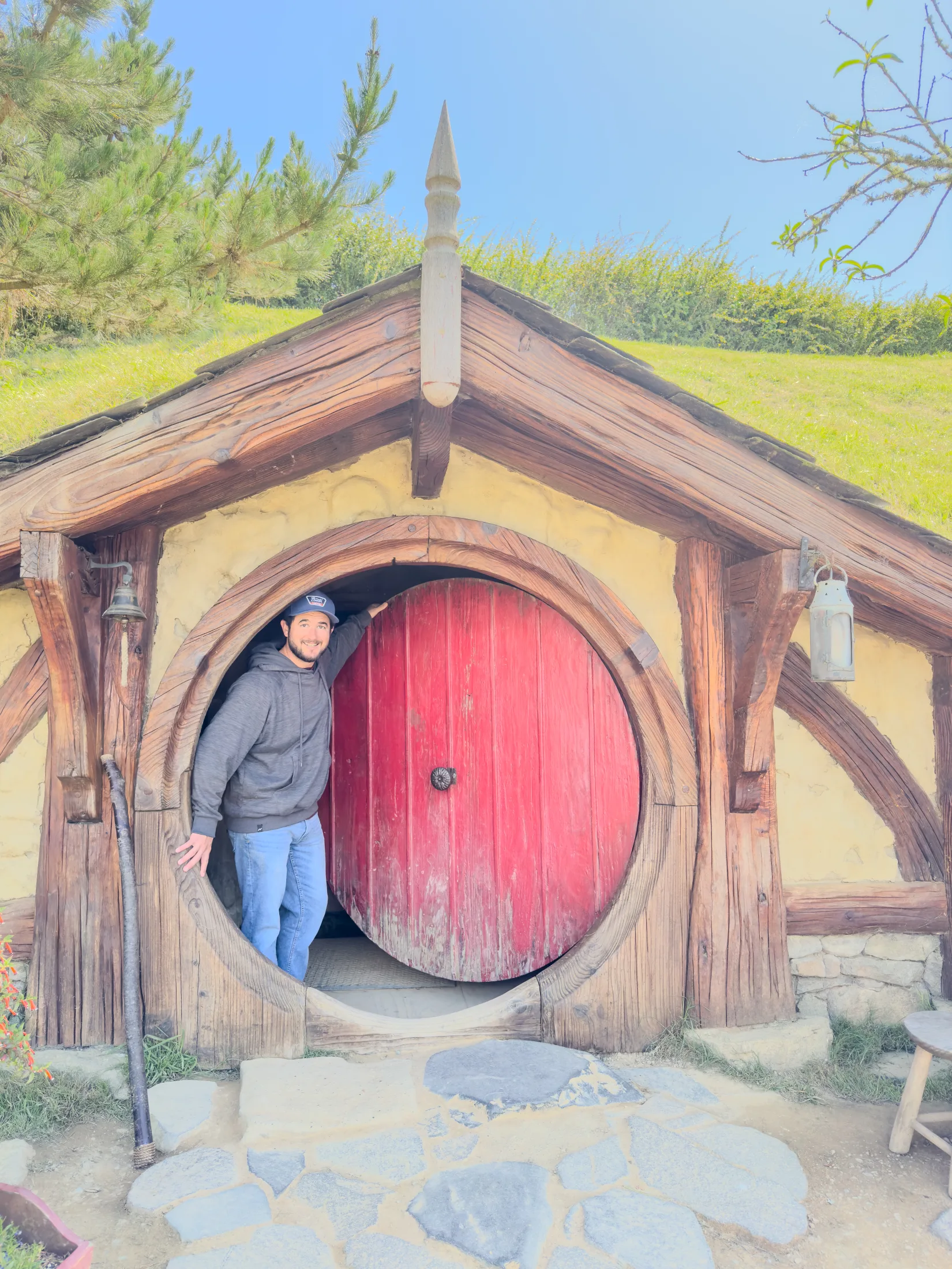 Alec standing in the round doorway of a hobbit hole with a red door and a grassy roof.