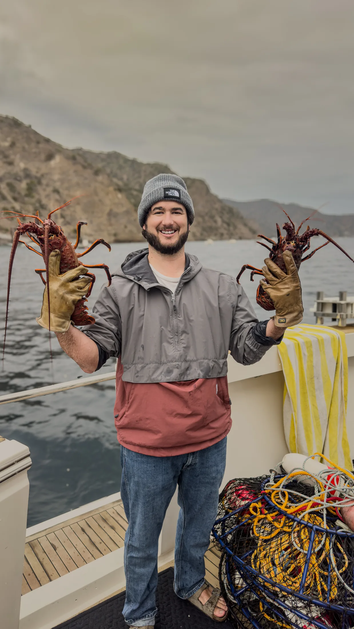 Alec on a boat deck in a beanie and rain jacket, holding a spiny lobster in each gloved hand, with traps and rope behind him.