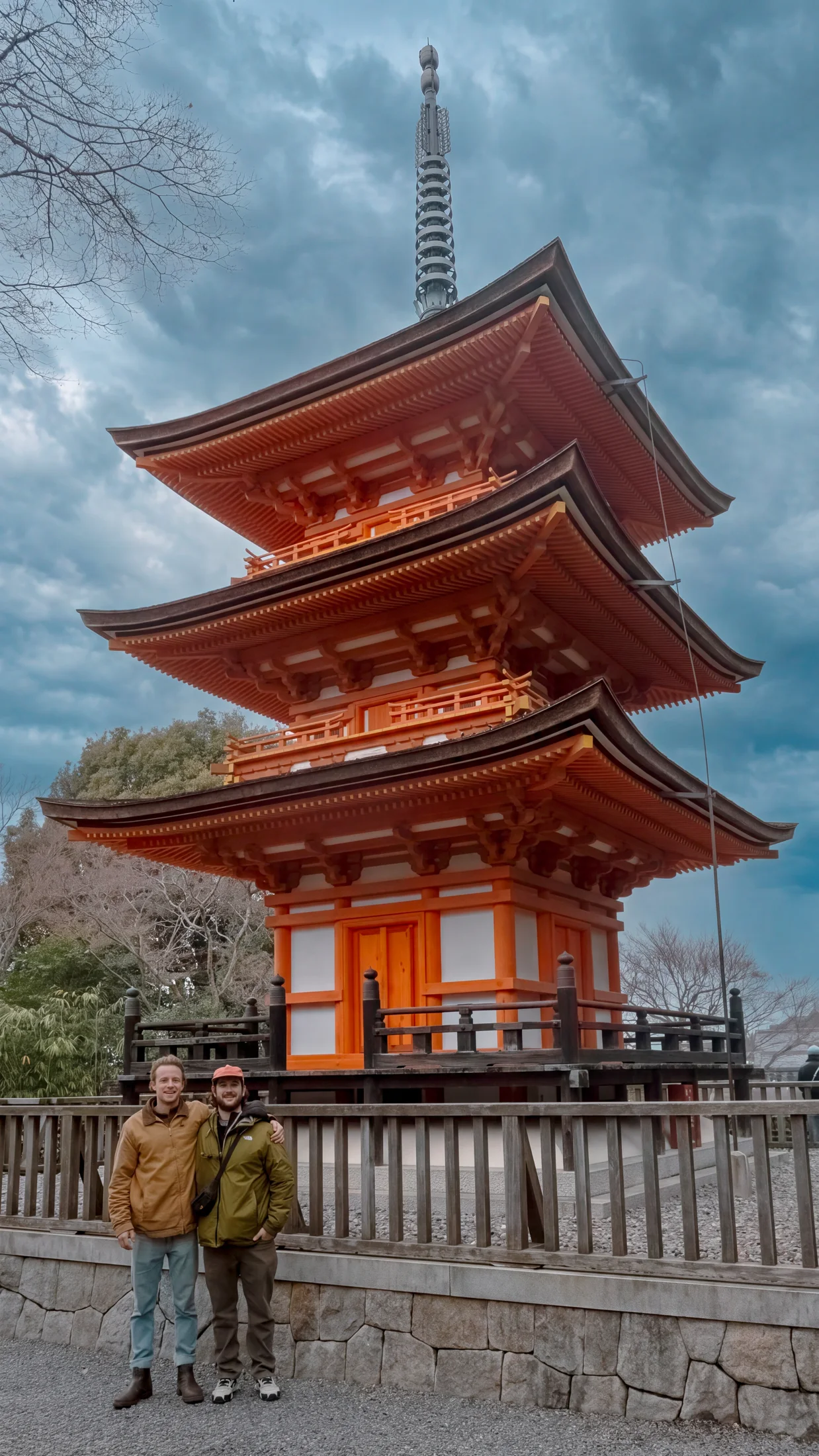 Two travelers standing beside a tall red pagoda beneath a cloudy sky.