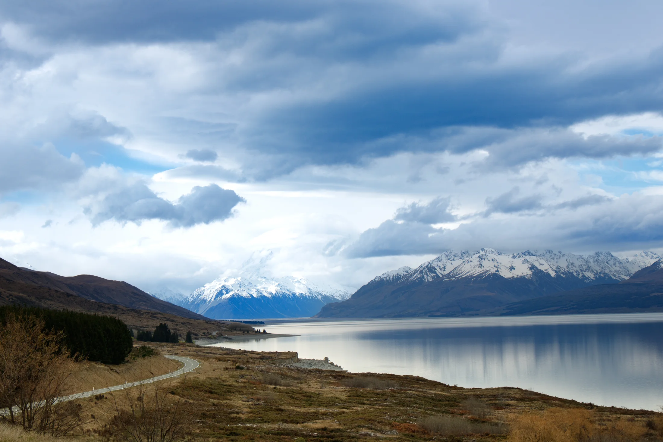 A winding road beside a still lake with snow-covered mountains beneath dark clouds.